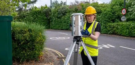 Dr Nat Easton setting up air pollution monitoring equipment (a metal cylinder on a stand) by the side of a road.