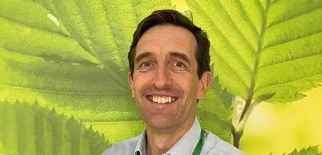 Dr Thomas Daniels standing in front of a wallpaper with large green leaves.