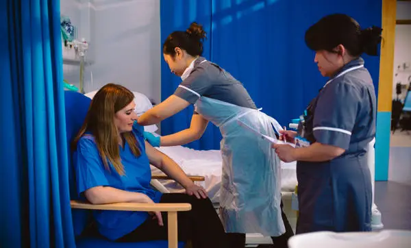 Healthcare staff assist a seated patient in a hospital ward beside a bed with blue privacy curtains.